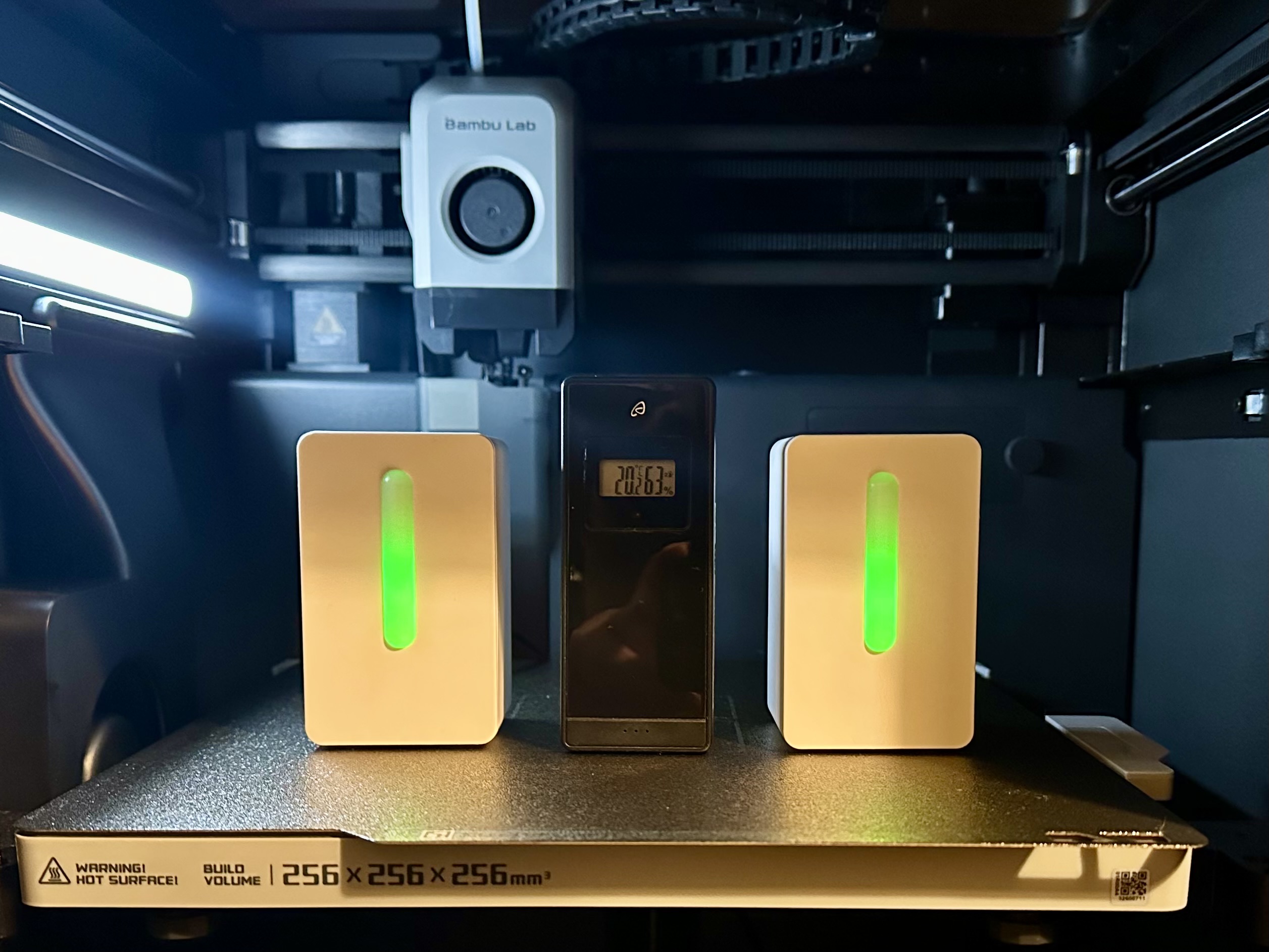 Photographs of 2 IKESP-Air air quality sensors flanking a "dumb" room temperature sensor on the bed of a Bambu Labs P1S 3D printer. The front LEDs on the IKESP-Air devices are green.