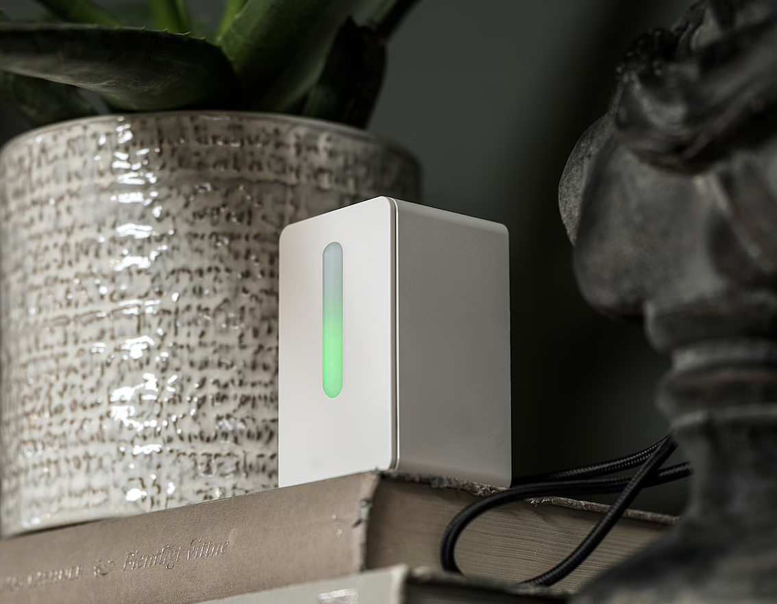 Image of an IKEA Vindriktning air quality sensor sitting on a book beside a potted plant. The LEDs on the front are glowing green.